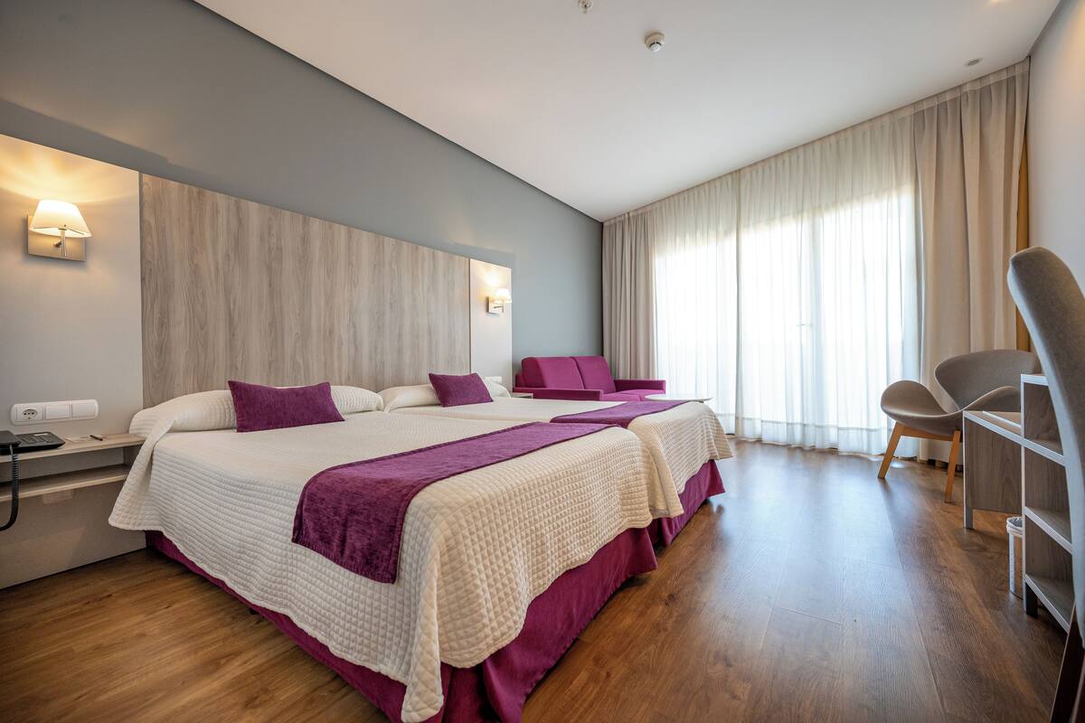 A hotel room with two beds, a desk, a chair, and a large window with curtains.