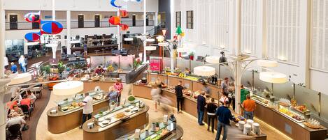 A spacious hotel lobby with multiple dining areas, a central bar, and a staircase leading to upper floors.