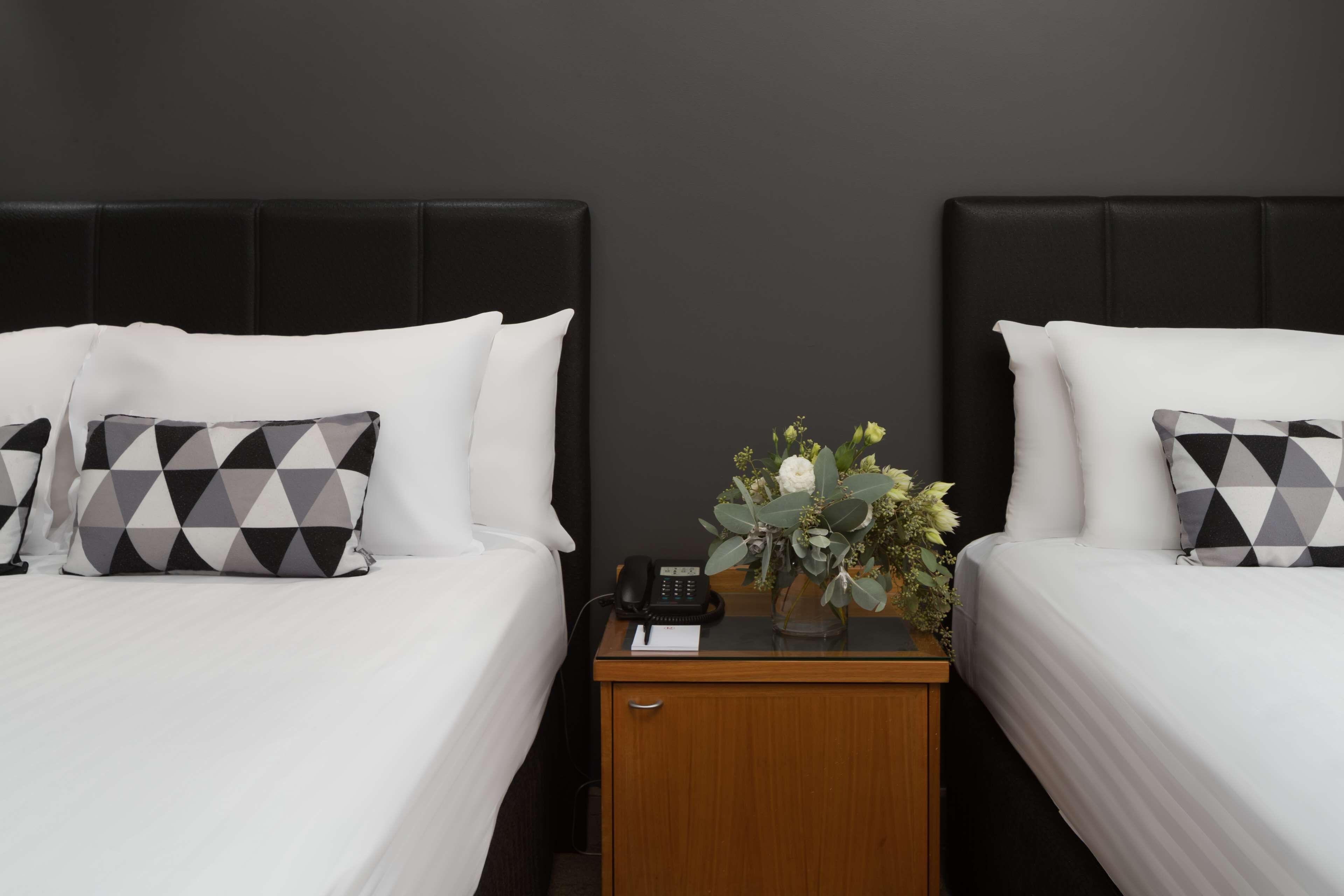 A hotel room with two beds, each with white bedding and patterned pillows. There is a wooden nightstand between the beds with a telephone and a vase with flowers.