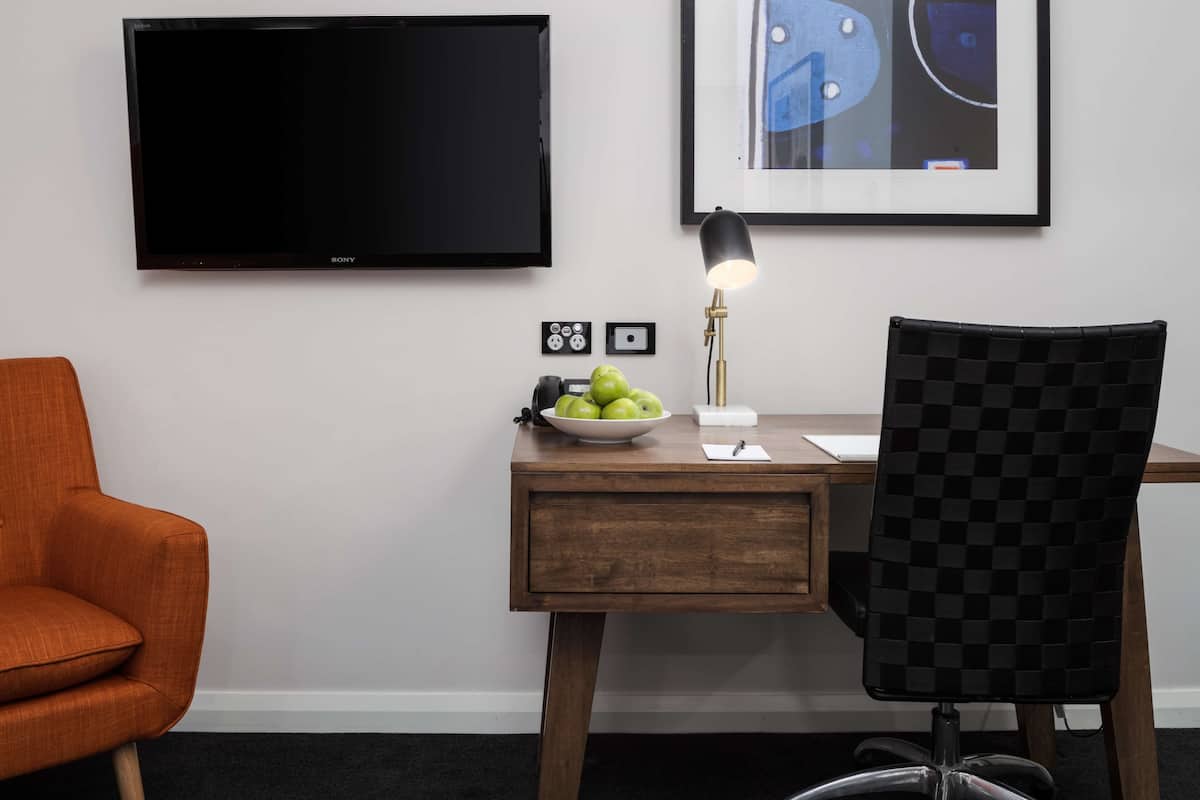A hotel room with a wooden desk, a black chair, a television, a framed artwork, and a bowl of green apples.