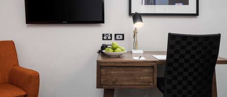 A hotel room with a wooden desk, a black chair, a television, a framed artwork, and a bowl of green apples.