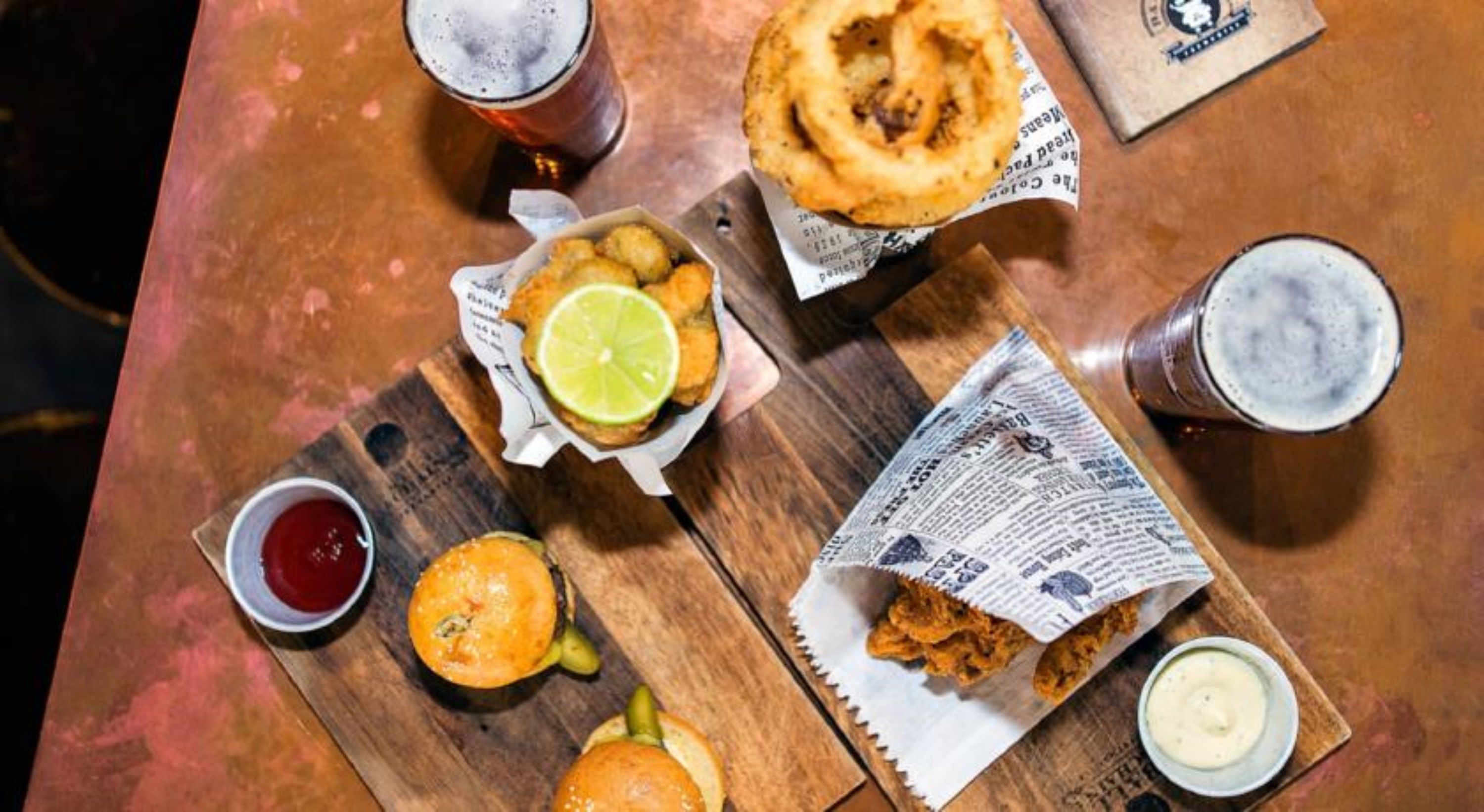 A meal consisting of fried fish, a burger, and a slice of pie, served on a wooden table with newspaper wrapping.