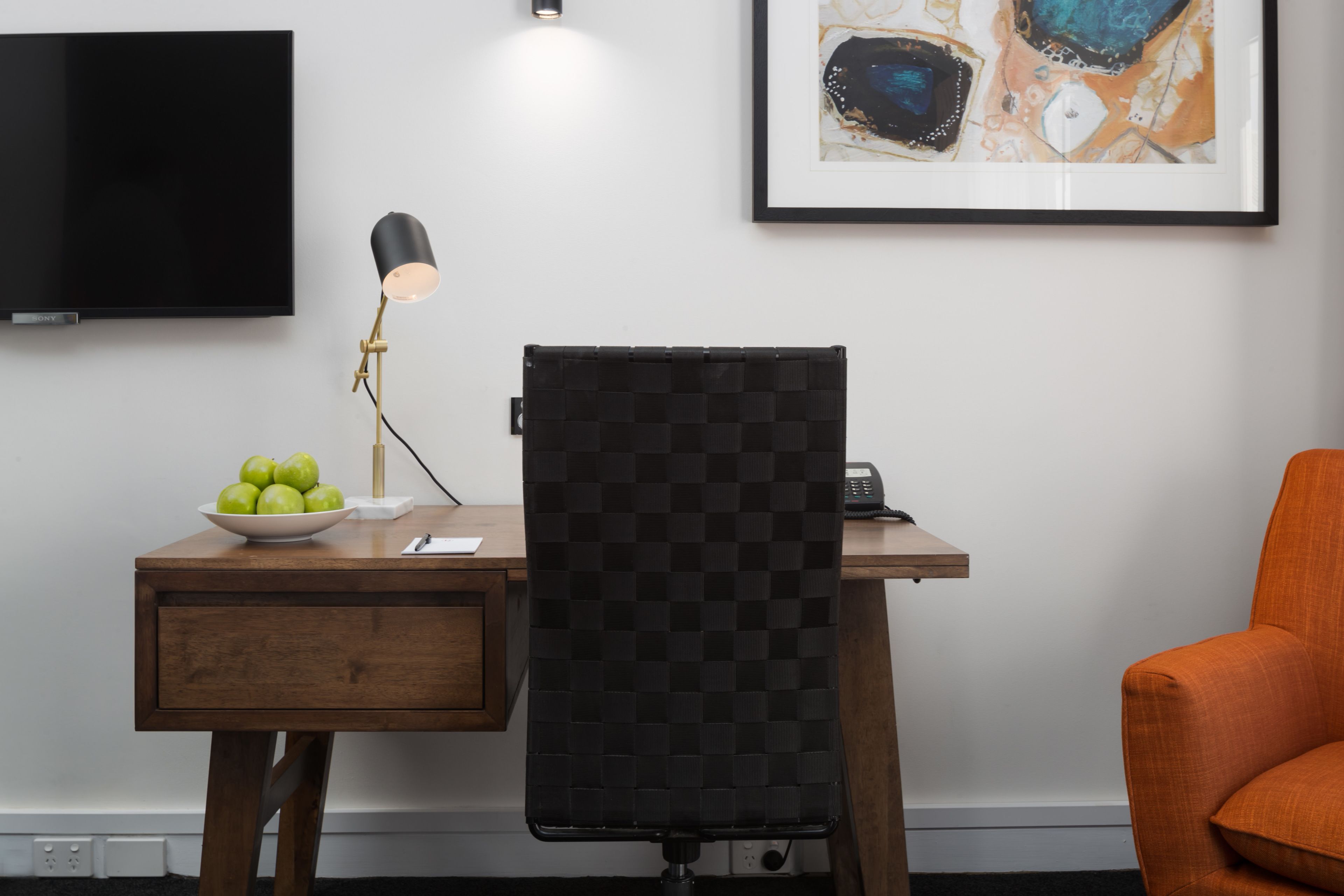 A modern office space with a wooden desk, a black office chair, a black lamp, a bowl of green apples, a framed abstract painting, and a flat-screen TV.