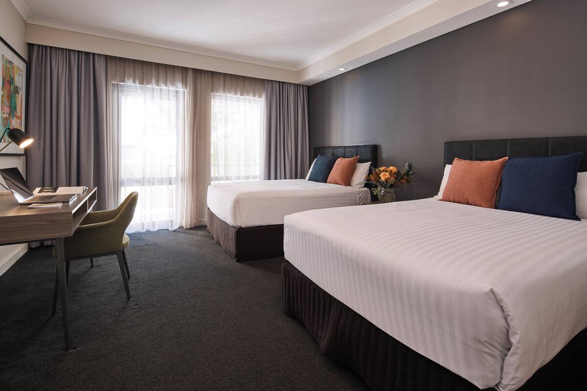 A hotel room with two beds, a desk with a chair, a lamp, and a large window with curtains.