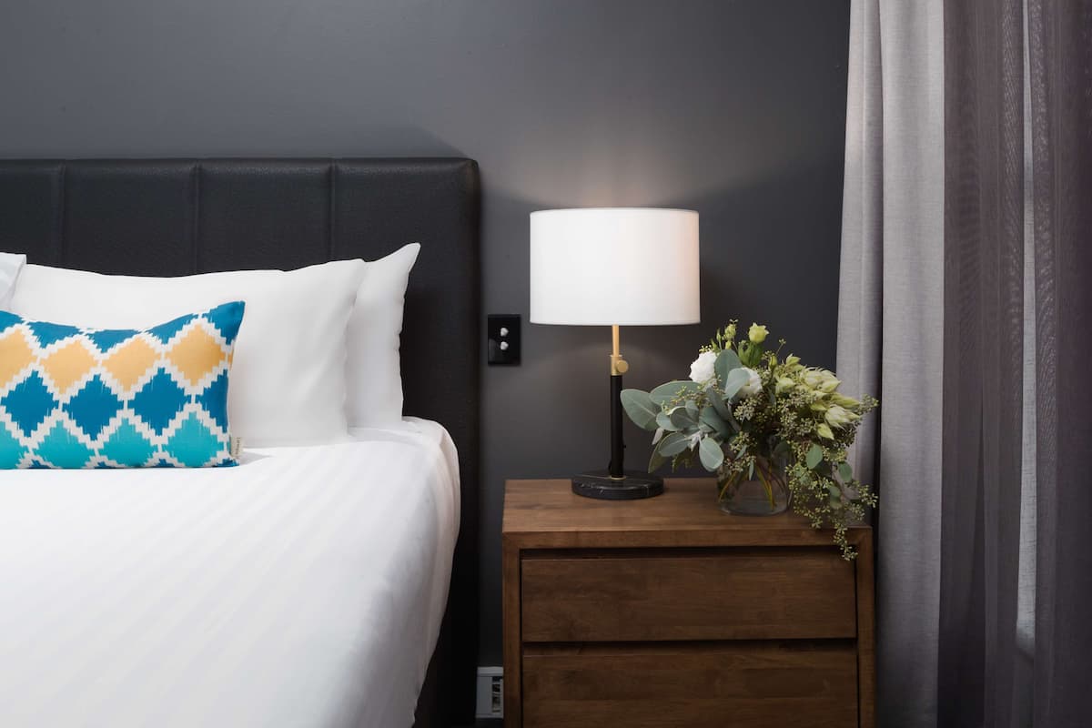 A neatly made bed with white and blue patterned pillows, a bedside lamp, and a vase with flowers on a wooden nightstand.