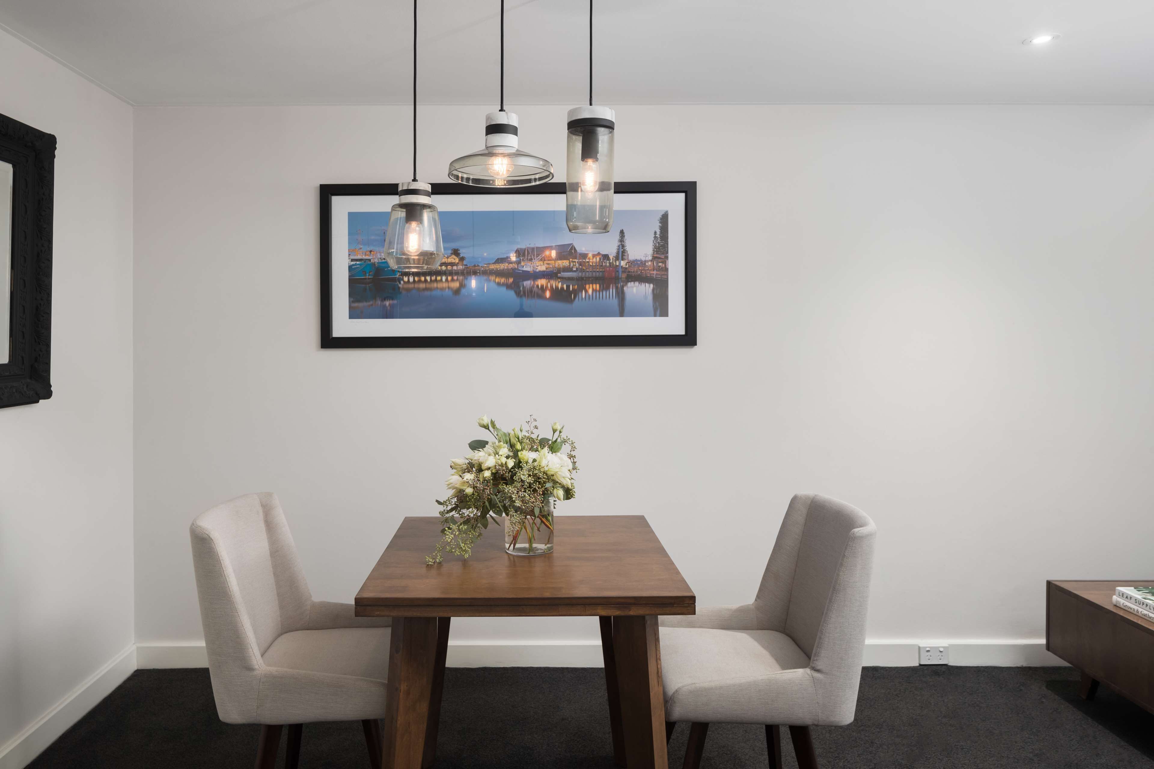 Un comedor con una mesa de madera, dos sillas, un jarrón de flores y un cuadro con una vista urbana enmarcada en la pared.