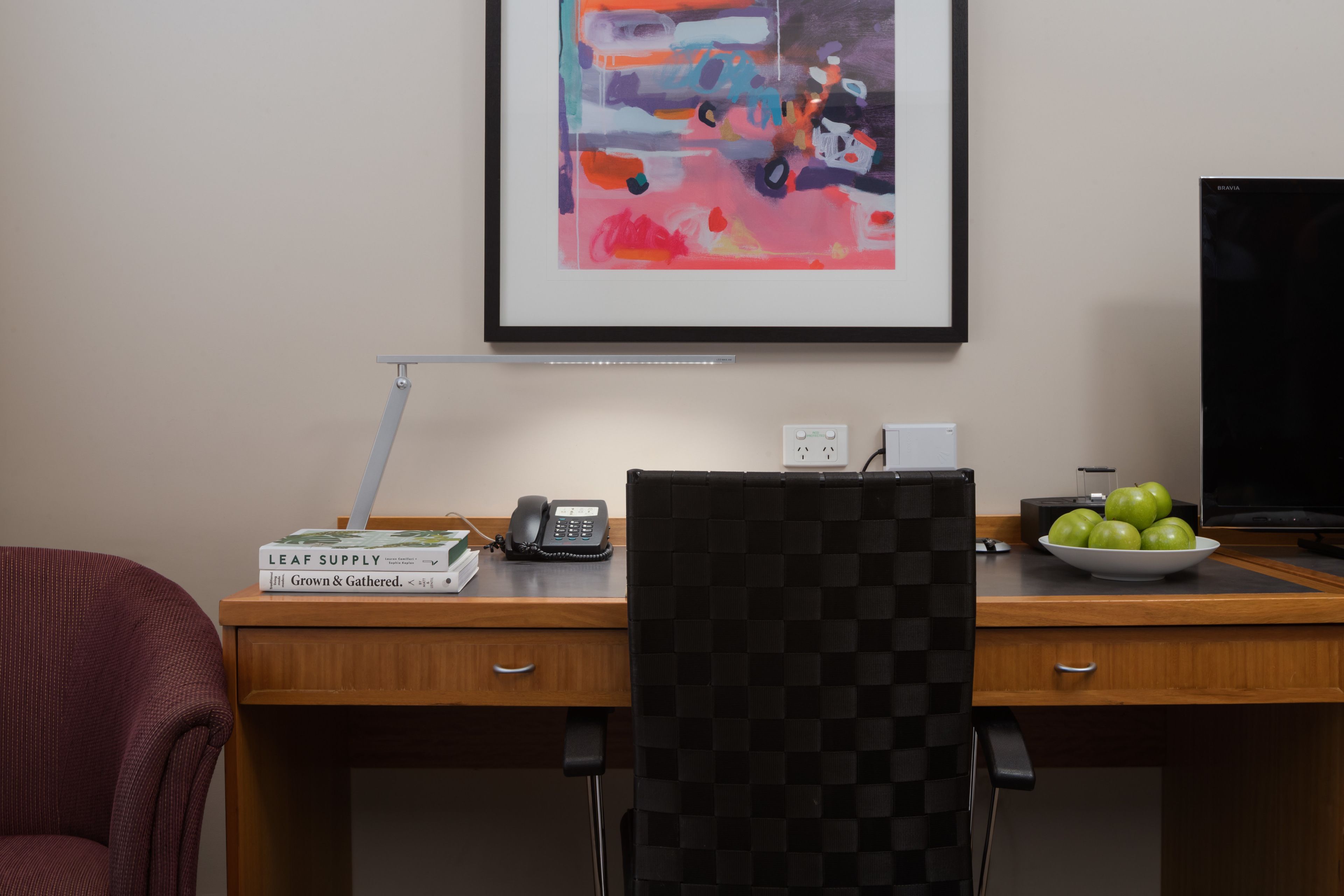 A wooden desk with a telephone, a bowl of green apples, and a framed abstract painting on the wall.