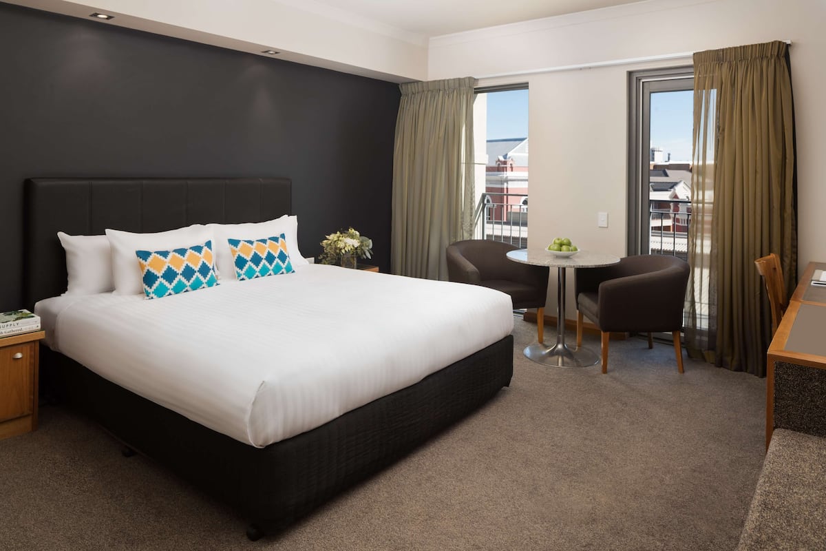 A hotel room with a large bed, a small table, two chairs, a nightstand, and a view of the city through the window.