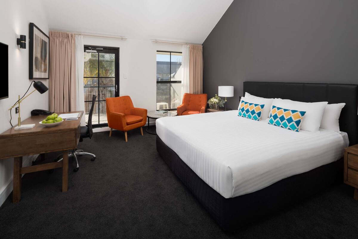 A hotel room with a large bed, two orange armchairs, a desk with a lamp, a chair, and a view of the outside through a sliding door.