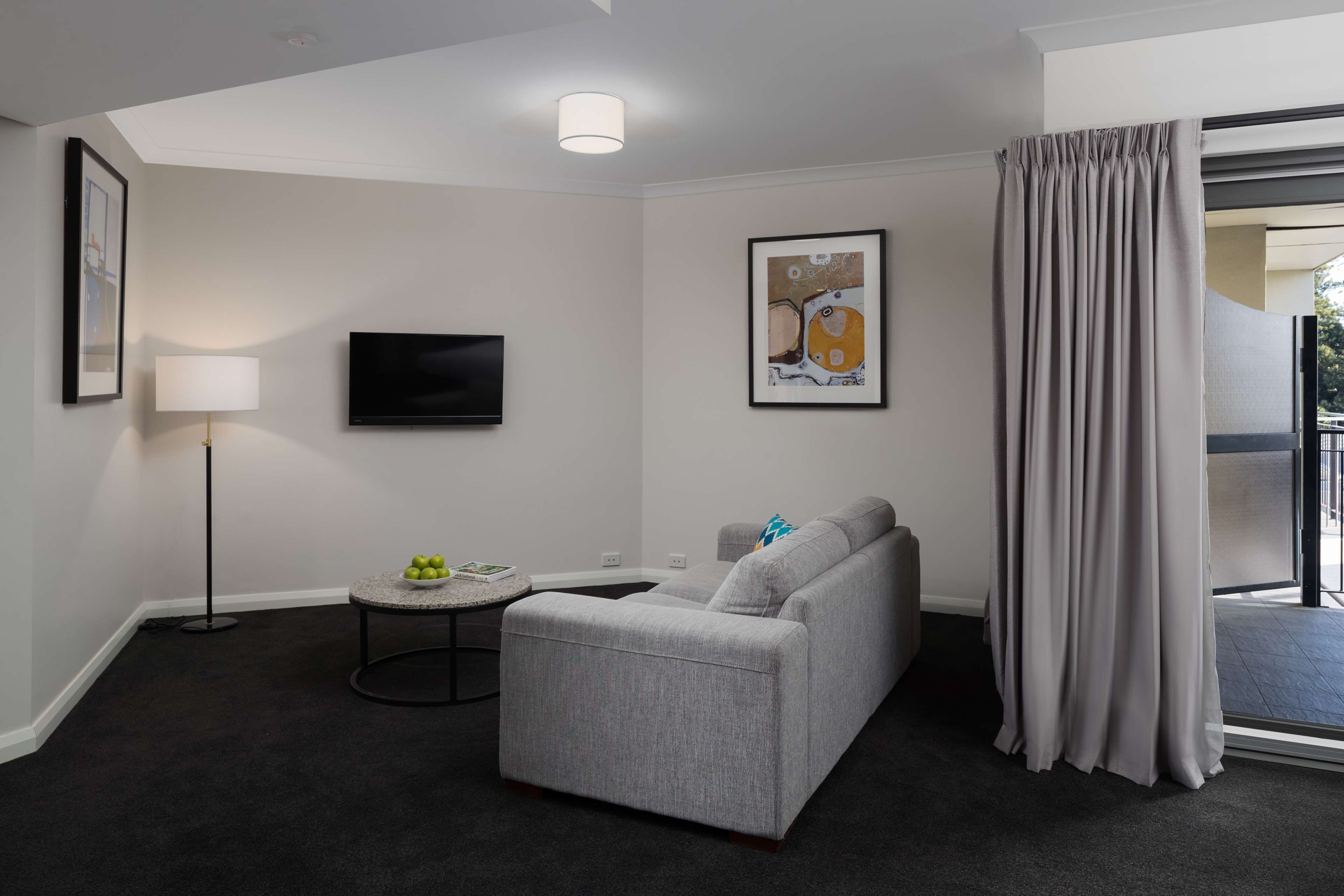 A modern hotel room with a grey sofa, a small round table with a lamp, a flat-screen TV, and a framed artwork on the wall.