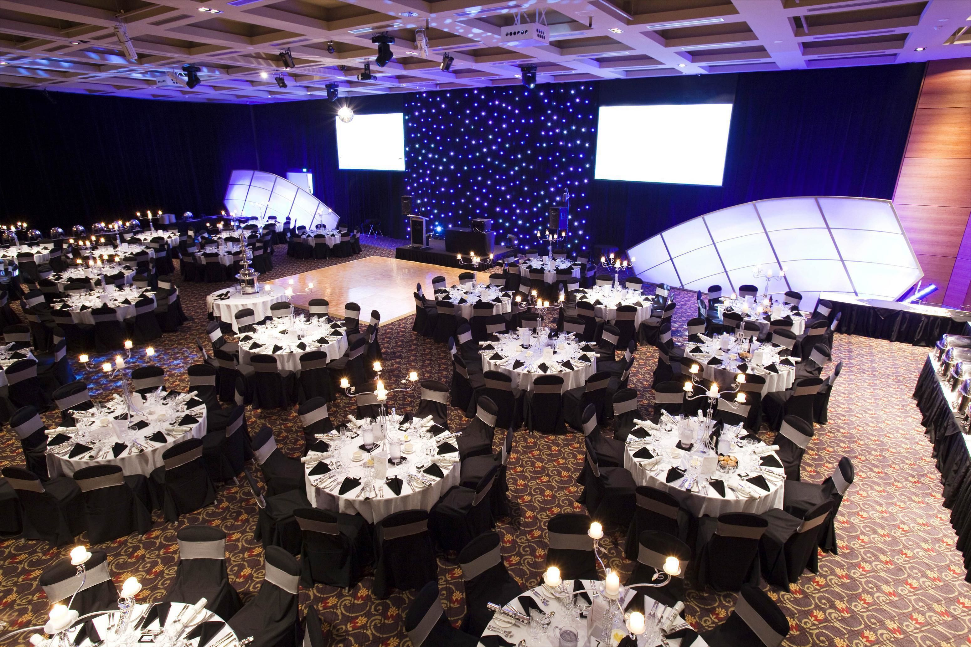 A large banquet hall with round tables set for a formal event, featuring a stage with a starry backdrop and large screens.