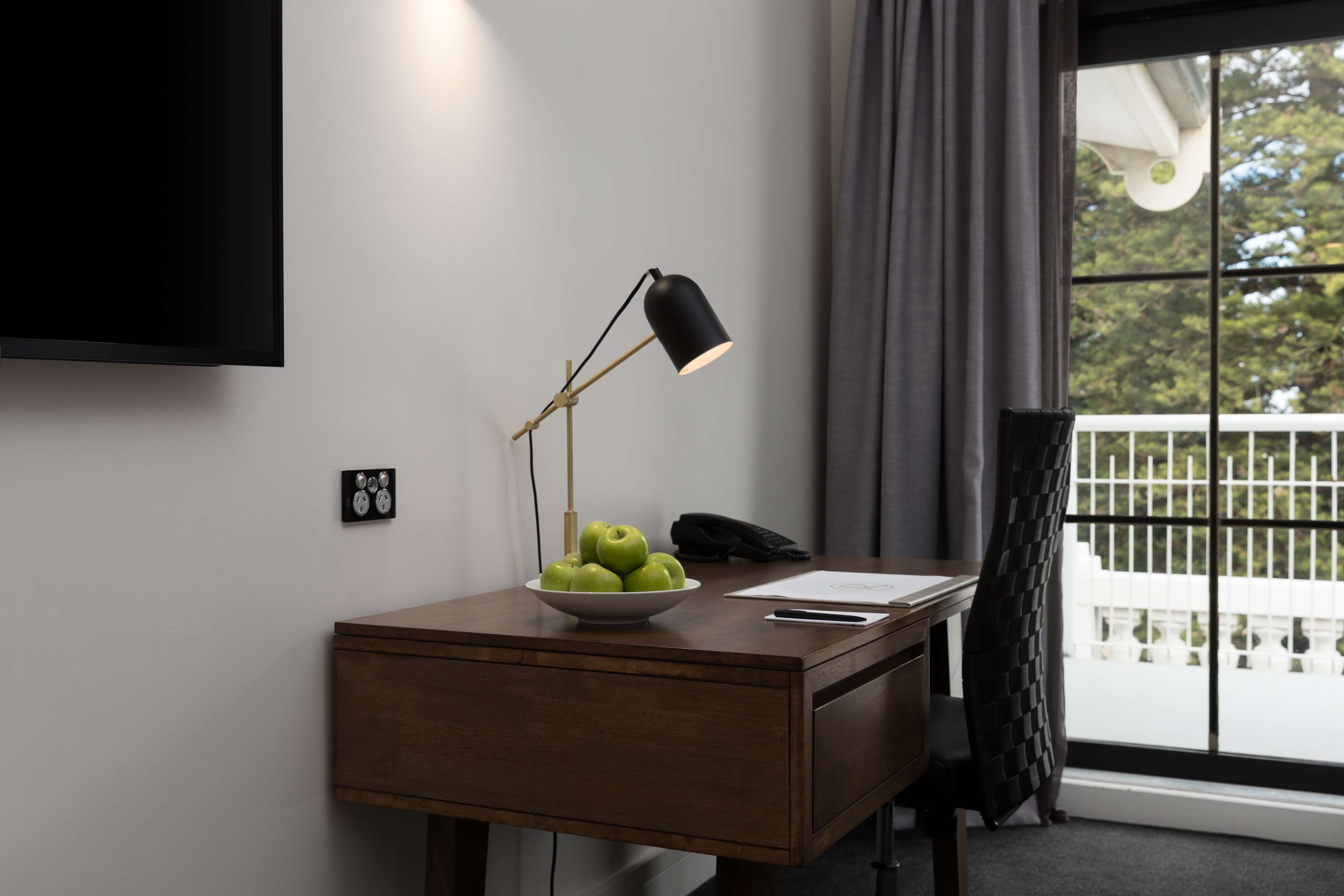 Habitación de hotel con un escritorio, un cuenco de manzanas verdes, una lámpara negra, un teléfono y un televisor montado en la pared.