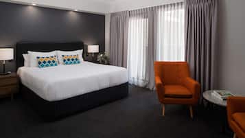 A hotel room with a large bed, a nightstand with a lamp, a chair, and a small table with a book.