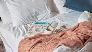 A bed with white bedding, a blue pillow, a pink knitted blanket, and a book titled "Breathe" with a floral design.