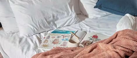 A bed with white bedding, a blue pillow, a pink knitted blanket, and a book titled "Breathe" with a floral design.