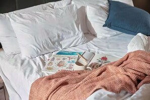 A bed with white bedding, a blue pillow, a pink knitted blanket, and a book titled "Breathe" with a floral design.