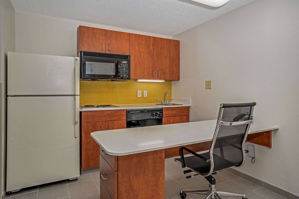 A compact kitchen with wooden cabinets, a microwave, a stove, and a built-in desk with a chair.