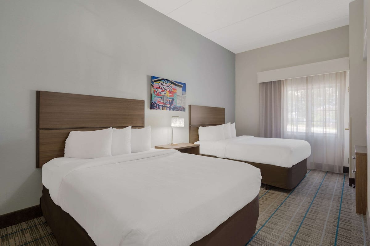 A hotel room with two beds, a nightstand, a lamp, a painting on the wall, and a window with curtains.