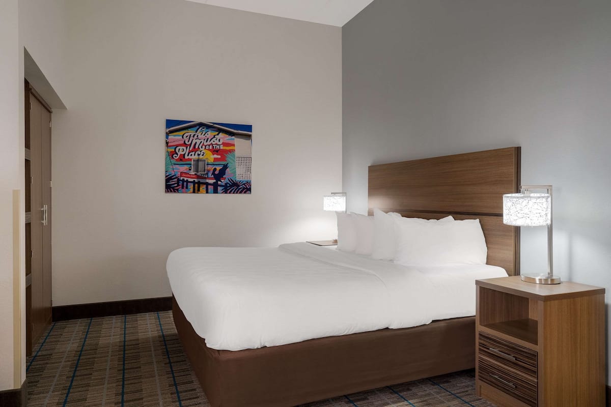 A hotel room with a large bed, a wooden headboard, a nightstand with a lamp, and a poster on the wall.