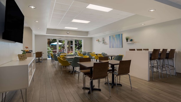 A modern dining area with wooden flooring, multiple tables and chairs, a white counter with stools, and a large flat-screen TV mounted on the wall. - MainStay Suites Wilmington - University Area (Wilmington)