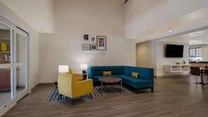 A modern hotel room with a blue sofa, a yellow armchair, a round coffee table, and a flat-screen TV mounted on the wall. - MainStay Suites Wilmington - University Area (Wilmington)