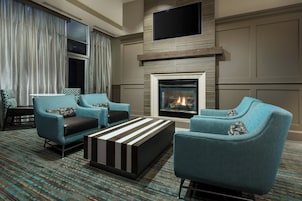A modern living room with two teal armchairs, a black and white striped ottoman, a fireplace, and a flat-screen TV mounted above the mantel.