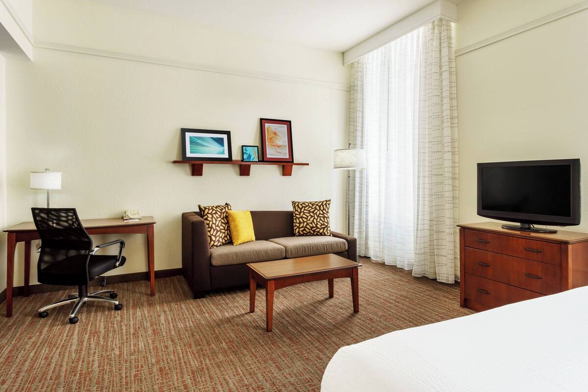 Ein Hotelzimmer mit Bett, Schreibtisch mit Stuhl, Sofa mit Kissen, Couchtisch, Fernseher auf einem Holzschrank und einem Fenster mit Vorhängen.