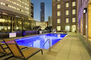An indoor swimming pool with a lifeguard warning sign, surrounded by modern buildings and a tall skyscraper in the background.