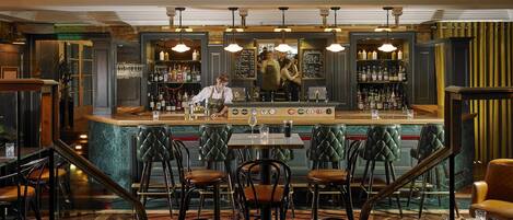 Zona de bar con un bartender, taburetes y una barra con diferentes botellas y vasos.