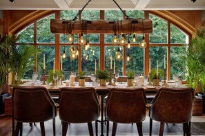 Comedor con una mesa grande dispuesta para cuatro personas, rodeada de sillas de madera, y vista a la vegetación a través de amplias ventanas.