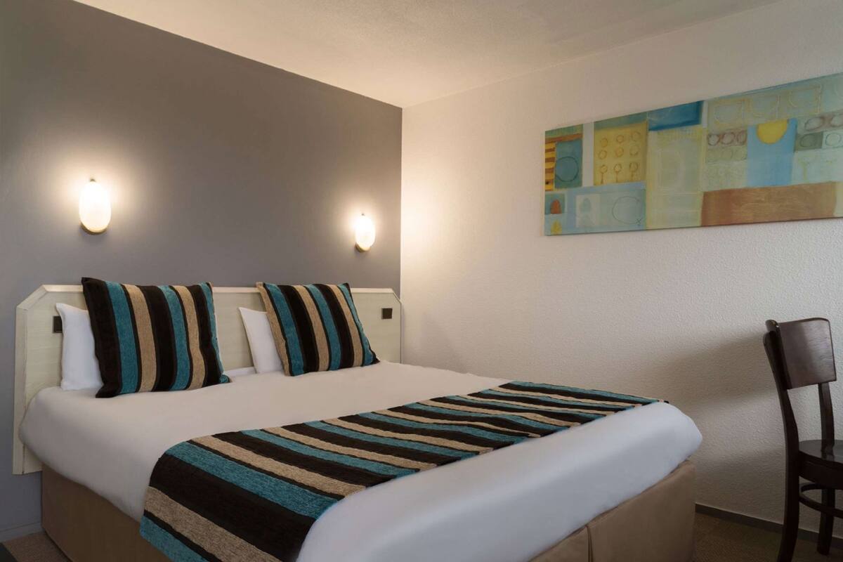 A hotel room with a bed, two wall-mounted lights, a chair, and a painting on the wall.