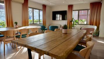 A conference room with a wooden table and chairs, large windows with curtains, and potted plants.