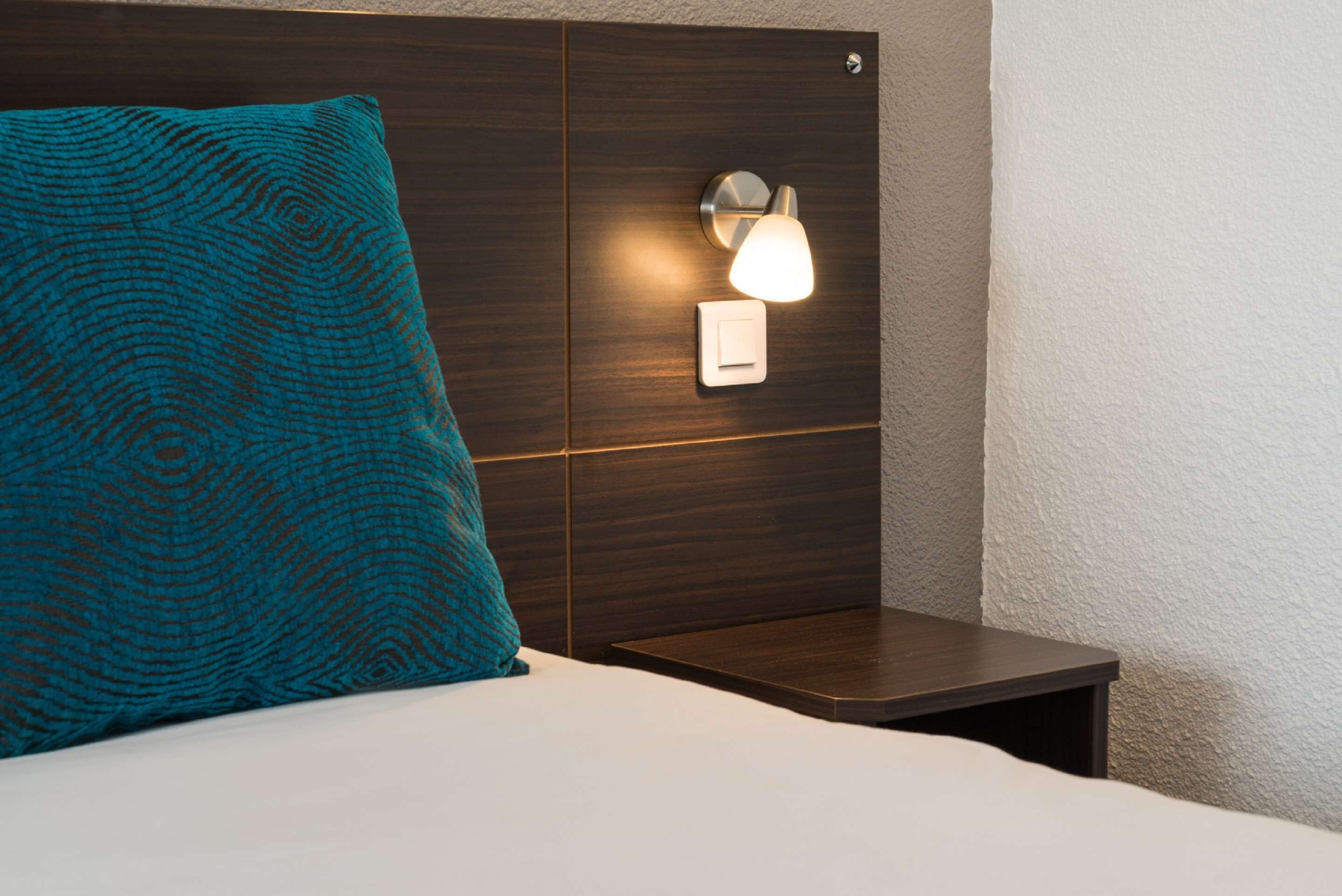 A neatly made bed with a teal patterned pillow, a bedside table, and a wall-mounted night light.