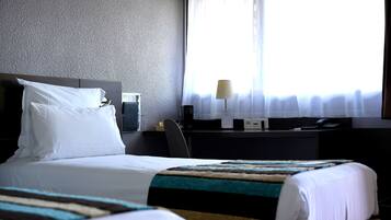 Ett hotellrum med en säng, ett skrivbord med en lampa och ett fönster med tunna, genomskinliga gardiner.