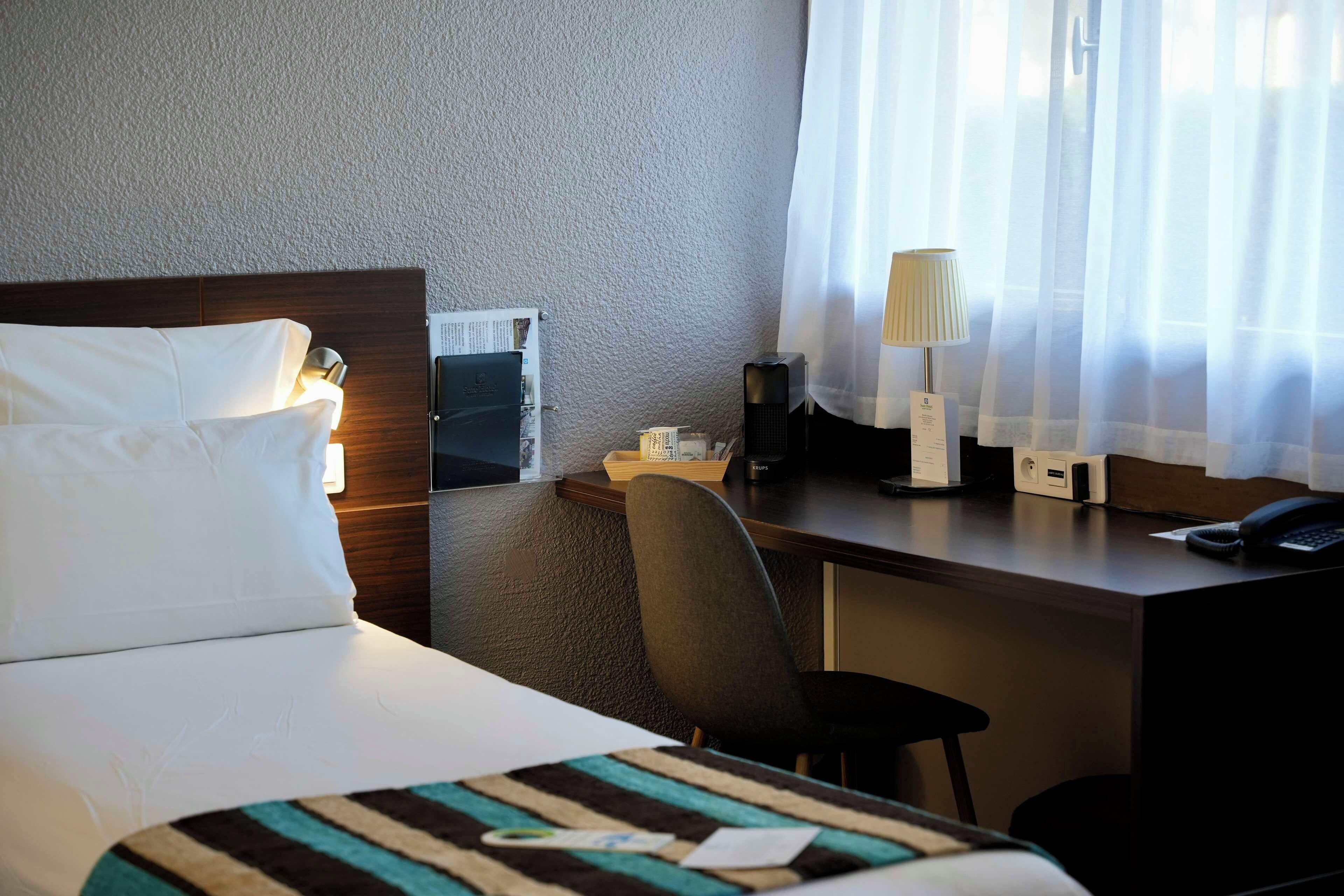 Une chambre d’hôtel comprenant un lit, un bureau, une chaise, une lampe, un téléphone et une fenêtre avec des rideaux légers.