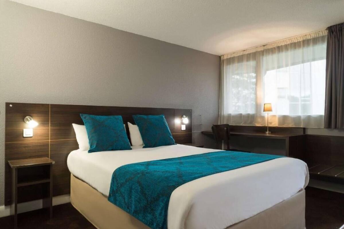 A hotel room with a bed, bedside table, lamp, and a view of the outdoors.