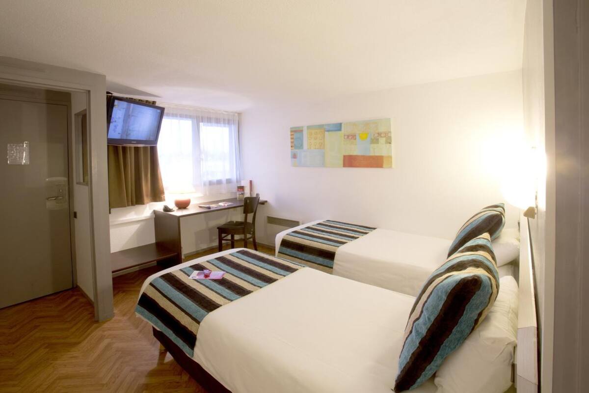 A hotel room with two beds, a desk, a television, and a window with curtains.