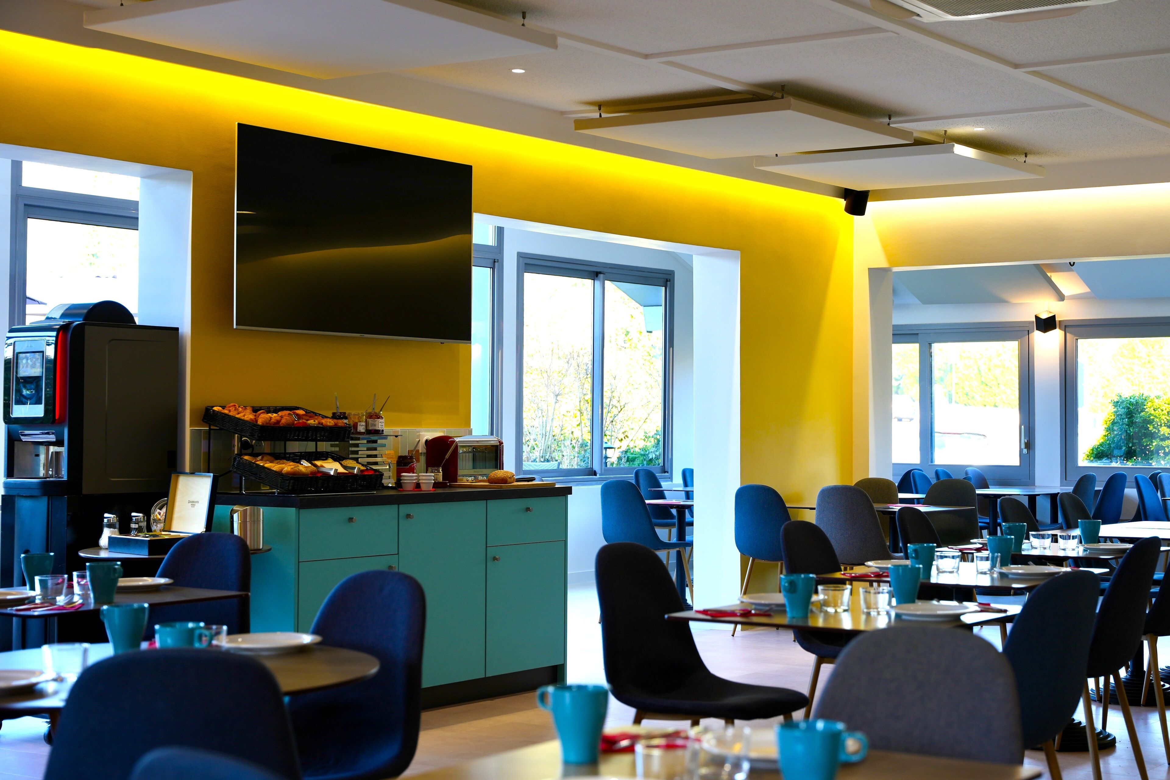 A modern dining area with yellow walls, blue chairs, and a large TV screen.