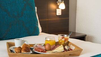 A breakfast tray with croissants, meats, cheese, a coffee cup, orange juice, and fruit on a bed in a hotel room.