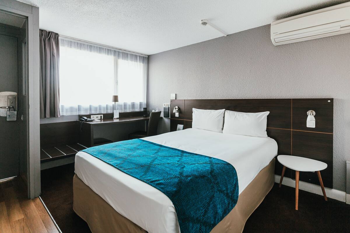 A hotel room with a large bed, a desk, a chair, a small table, and a window with curtains.