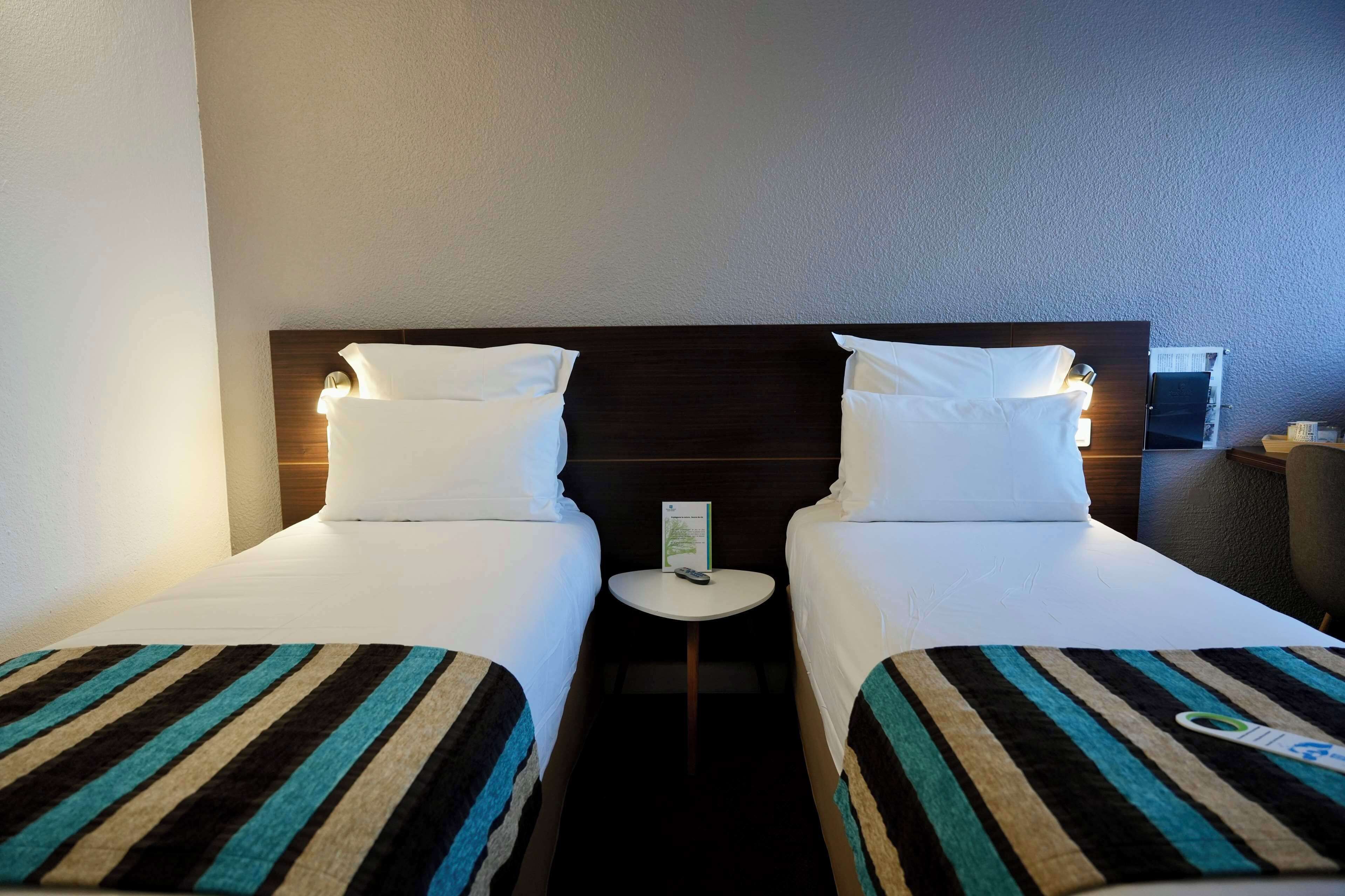A hotel room with two single beds, a small bedside table with a remote control, and a wall-mounted air conditioning unit.