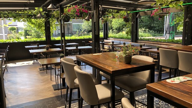 An outdoor cafe with wooden tables and chairs, hanging plants, and a view of greenery through large windows. - Hotel Chariot (Aalsmeer)