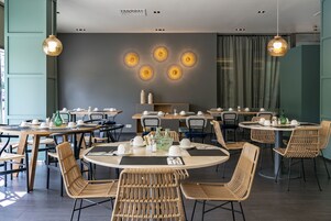 A modern dining area with round tables and wicker chairs, featuring a central wall with circular light fixtures and a ceiling with recessed lighting.