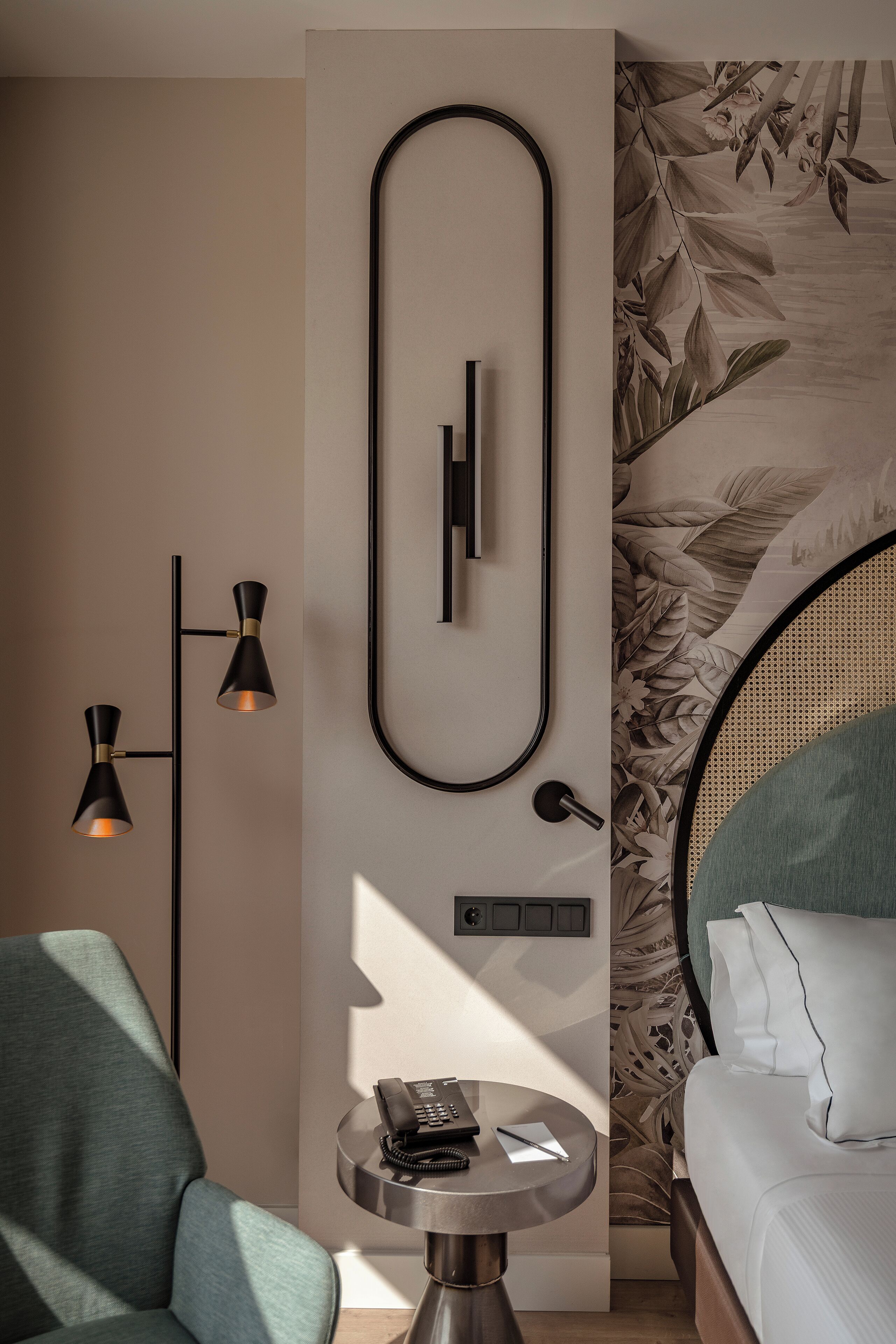 Una habitación de hotel moderna con una cama, una mesita redonda con teléfono y bloc de notas, una lámpara de pared y una pared decorada con un estampado floral.