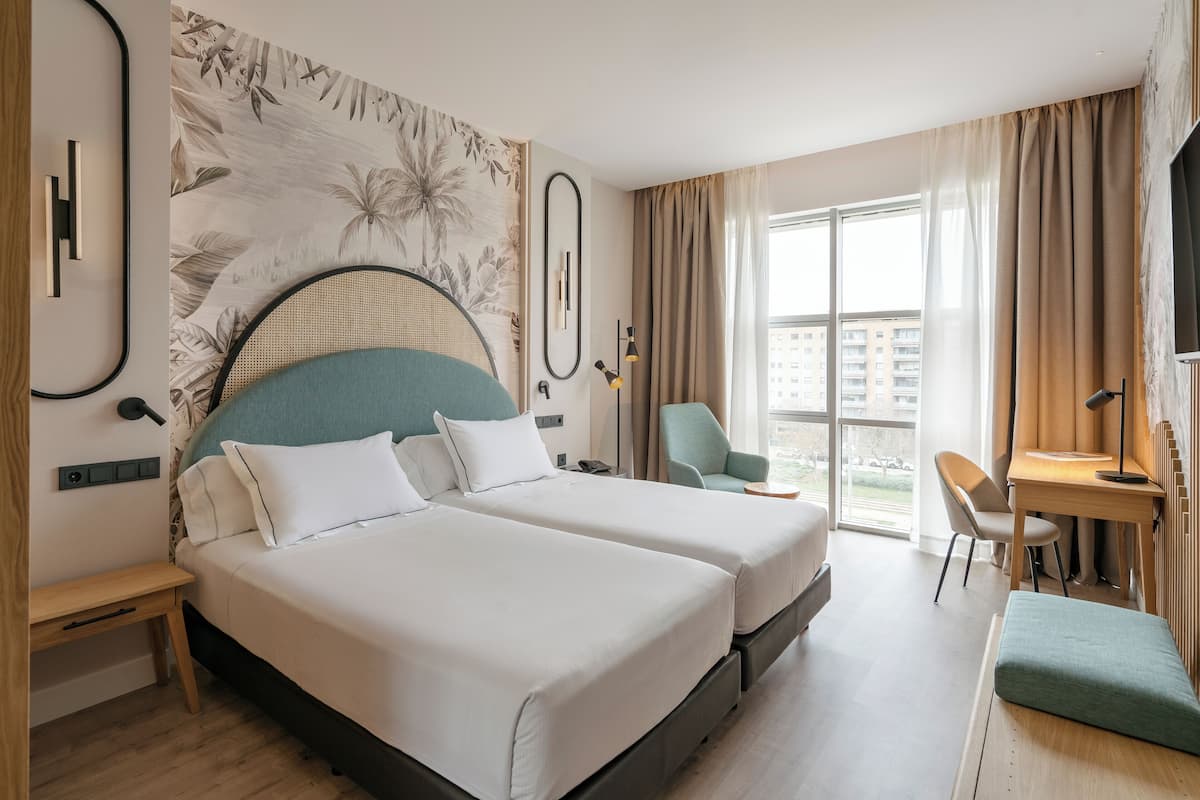 Hotellihuone, jossa on kaksi sänkyä, työpöytä, tuoli ja suuri ikkuna, jossa on verhot.