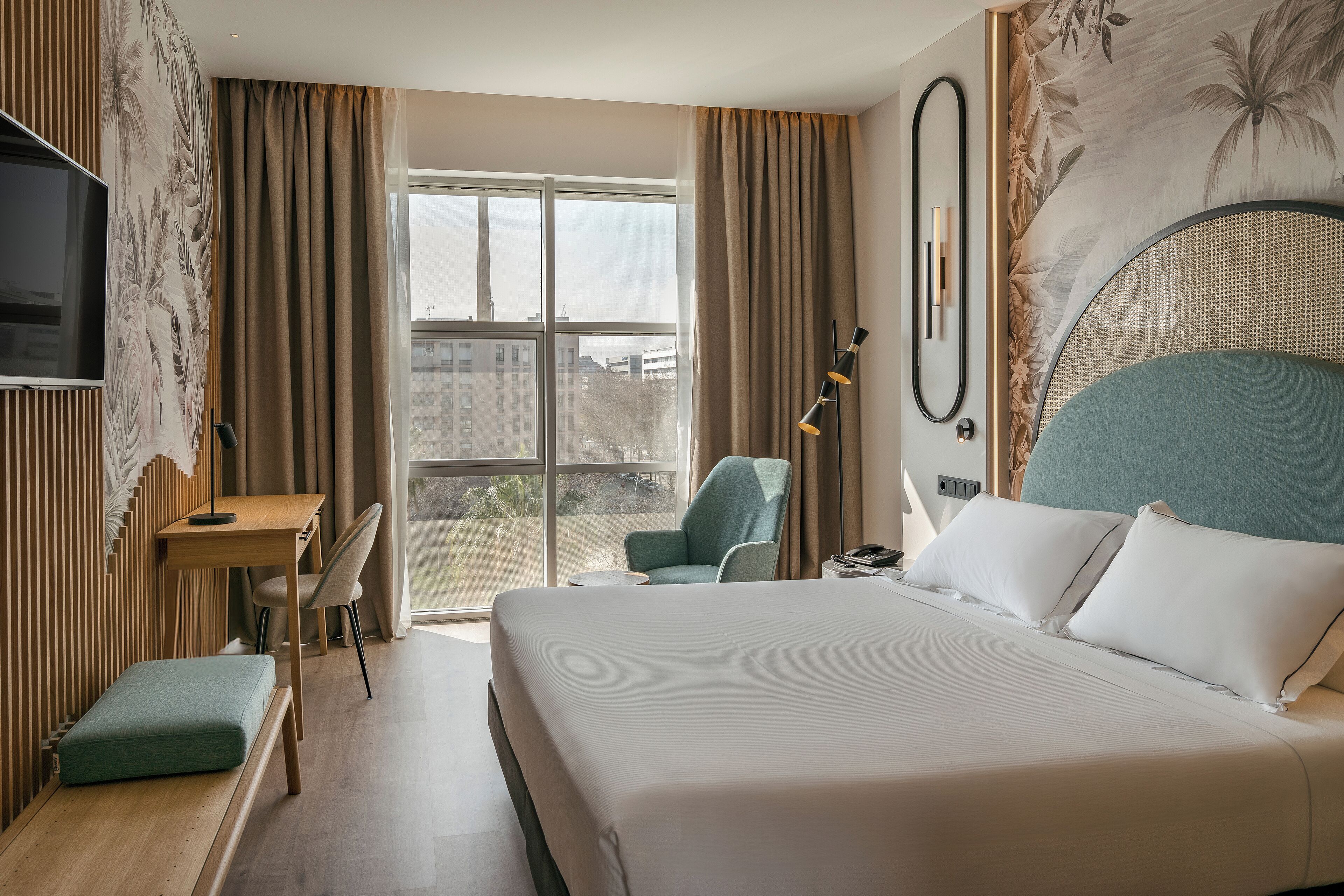 A hotel room with a large bed, a desk, a chair, a TV, and a view of the city.
