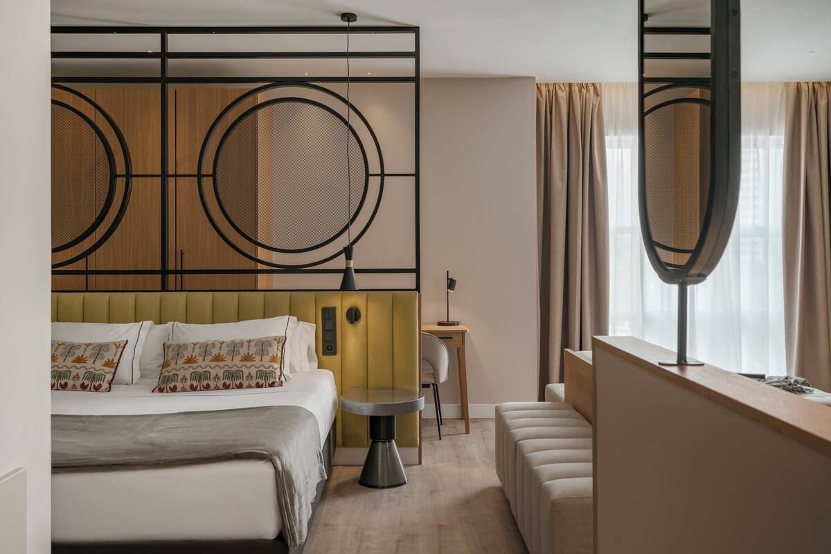 Hotellihuoneessa on sänky, pieni pyöreä pöytä, työpöytä ja suuri ikkuna, jossa on verhot.