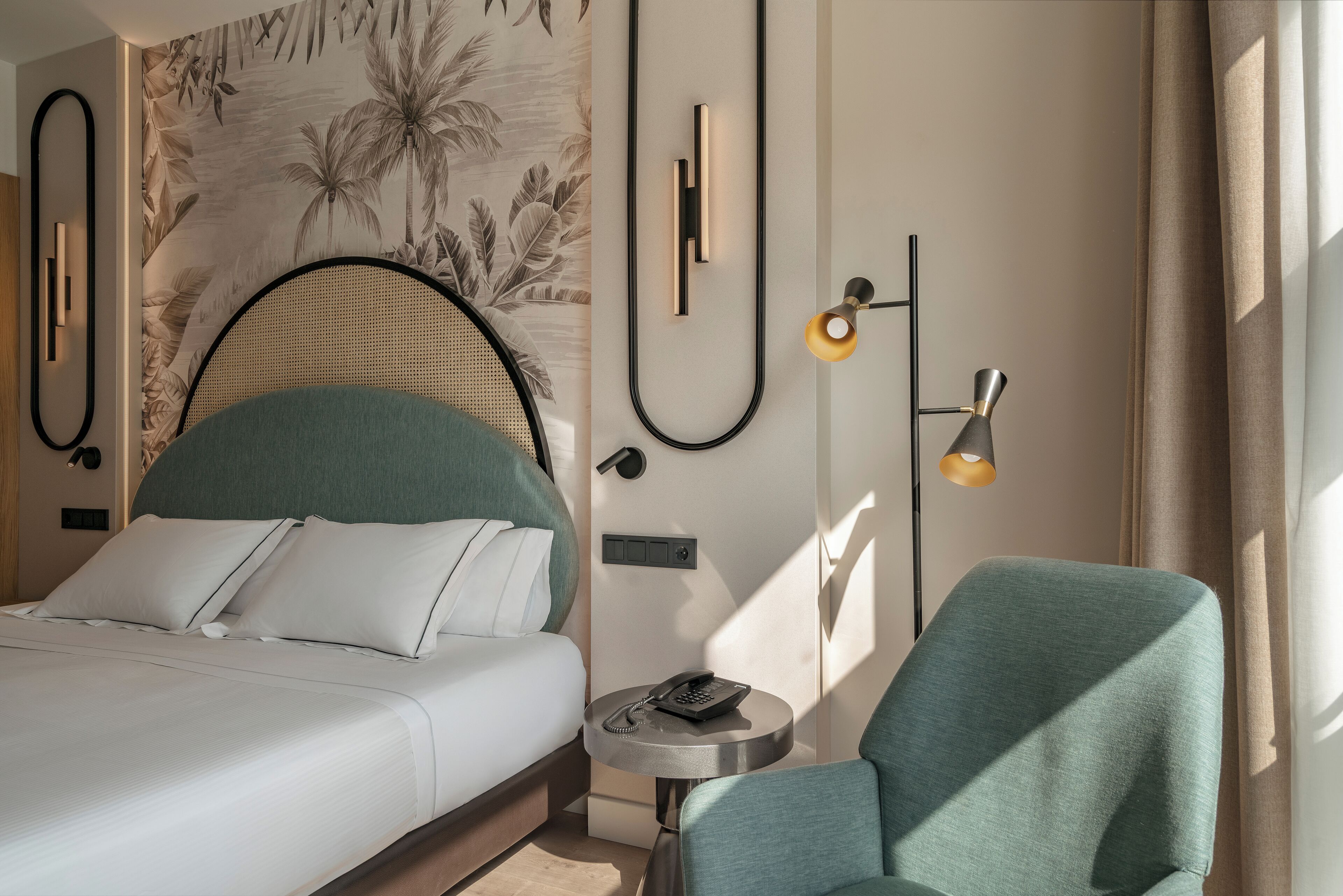A hotel room with a bed, a round table with a telephone, a chair, a wall-mounted lamp, and a tropical wallpaper.
