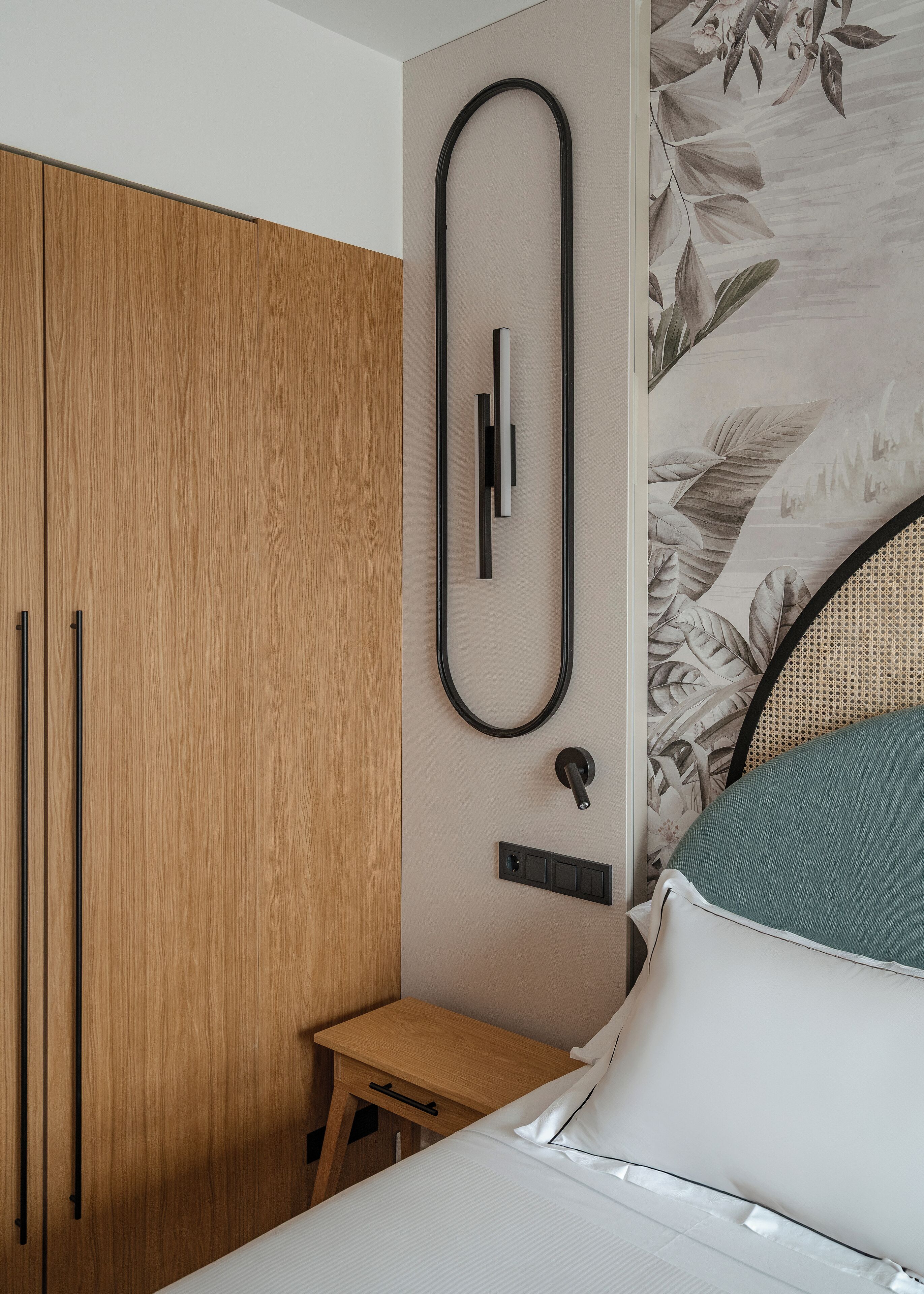 Un dormitorio con un armario de madera, una mesita de noche con cajón, una cama con ropa de cama blanca y una lámpara fijada a la pared.