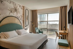 A modern hotel room with a large bed, a desk, a chair, and a view of the city.